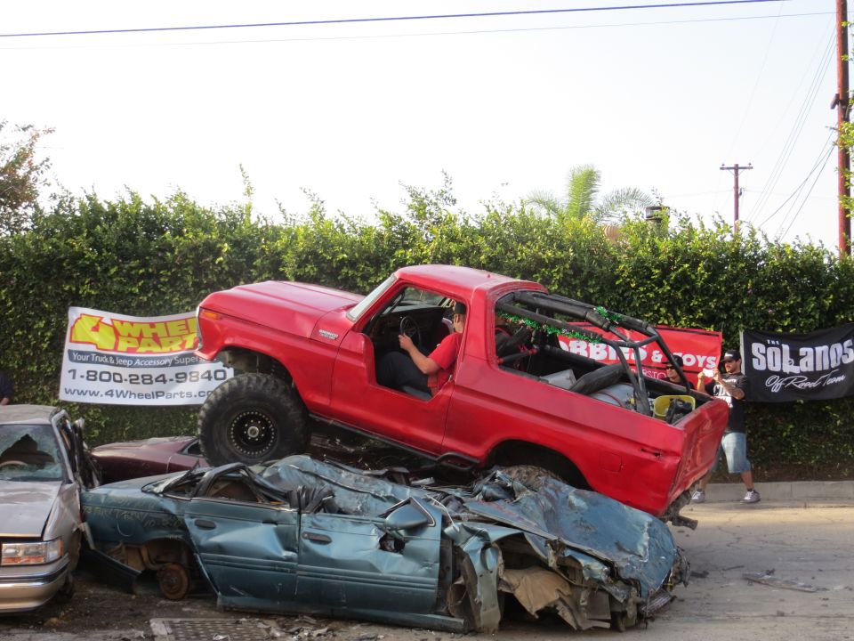 Red 1978 Ford Bronco on top of two crushed cars, with a "4WheelParts.com" banner in the background