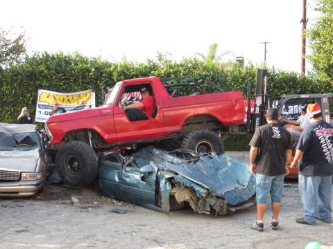 1978 Ford Bronco - A red lifted truck is positioned on top of a heavily damaged blue car, with onlookers present.