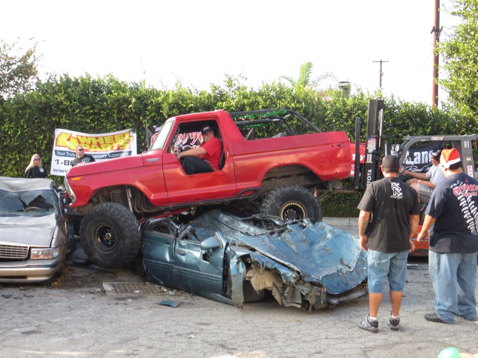 Red lifted Ford Bronco on top of a crushed blue sedan. People watching