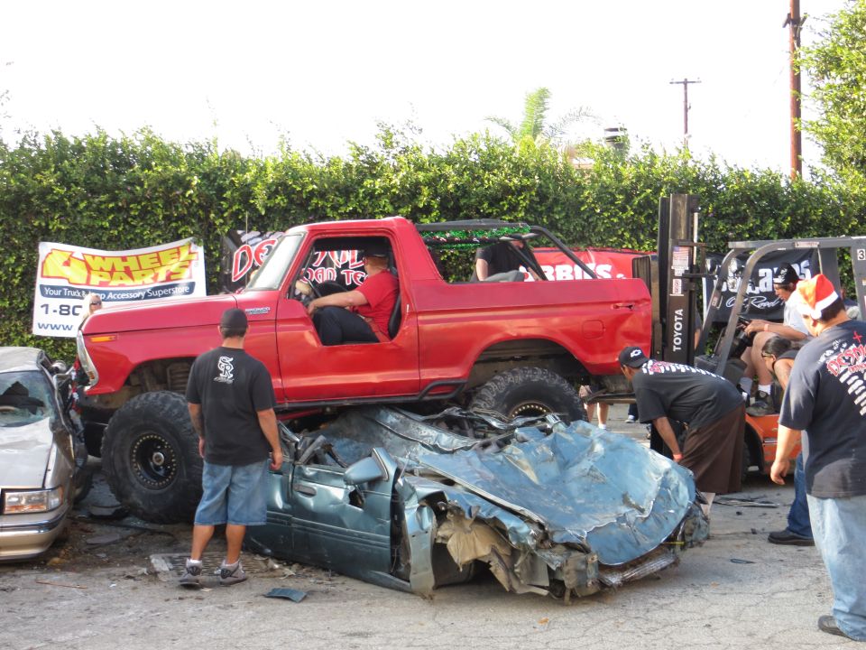 Red 1978 Ford Bronco crushing a blue car, lifted on large tires