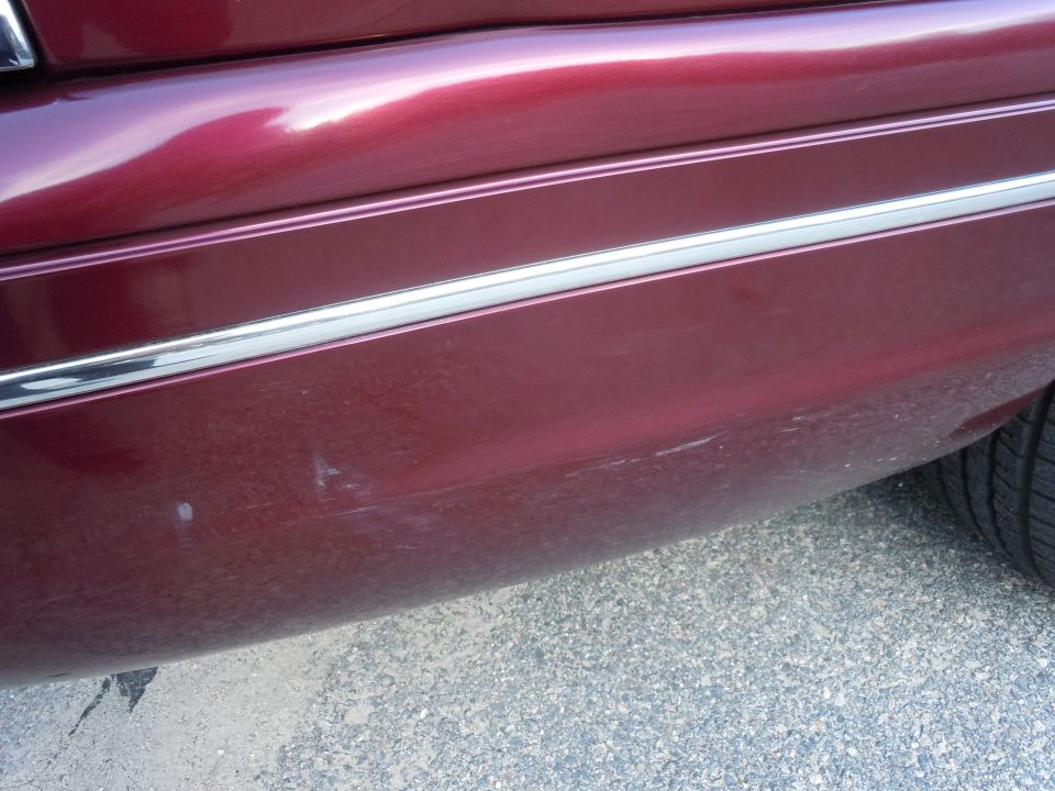 Maroon car rear bumper corner with chrome trim and scratches. Tire visible on right