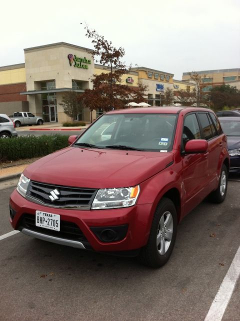 1987 Suzuki Samurai - This red Suzuki SUV is parked in a lot with a shopping center in the background.