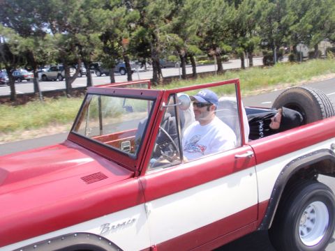 1968 Ford Bronco - Driver-side view of a topless red-and-white early Bronco being driven outdoors, with a man in a blue ball cap, sunglasses and white graphic…