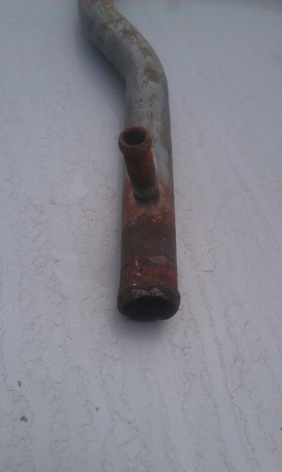 Rusty metal radiator hose with a smaller attached pipe, lying on a textured surface