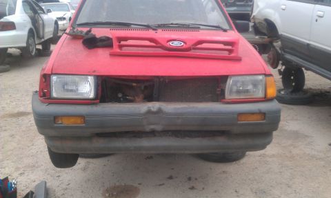 1991 Ford Festiva - A red 1991 Ford Festiva sits in a junkyard with its grille removed and other vehicles in the background.