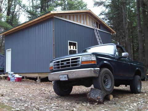 1995 Ford Bronco - A dark pickup truck is parked in front of a blue building with a ladder leaning against it.