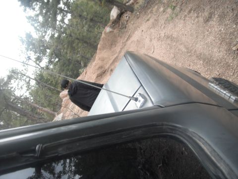1990 Ford Bronco - A gray SUV, likely a 1990 Ford Bronco, is seen from a high angle on a dirt road with trees and rocks in the background.