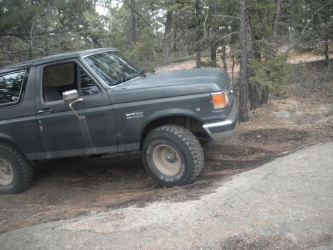 1990 Ford Bronco - A dark gray Ford Bronco from the late 1980s is seen off-road in a wooded area.