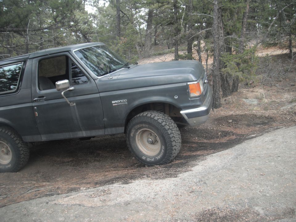Dark gray Ford Bronco, 1987 model year, parked on a dirt road with trees in the background