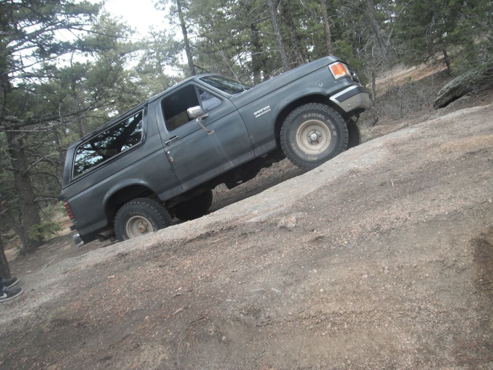 1990 Ford Bronco climbing a steep dirt incline with trees in the background