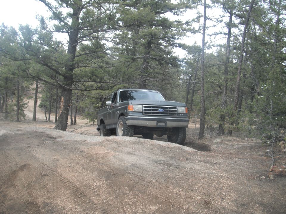 Green Ford Bronco on dirt road, front 3/4 view, trees in background