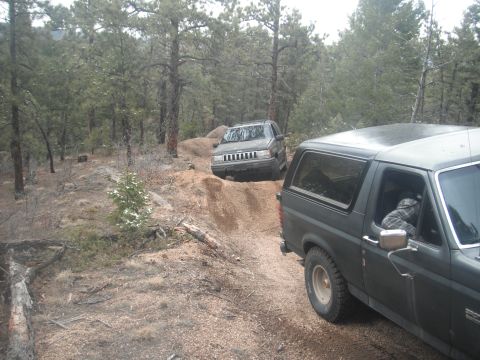 1990 Ford Bronco - Two SUVs navigate a wooded trail, with the Ford Bronco in the foreground and the Jeep Grand Cherokee climbing a hill.