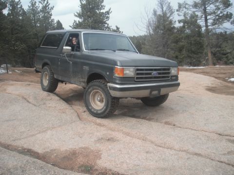 1990 Ford Bronco - A 1990 Ford Bronco is shown off-roading on a rocky path in a wooded area.