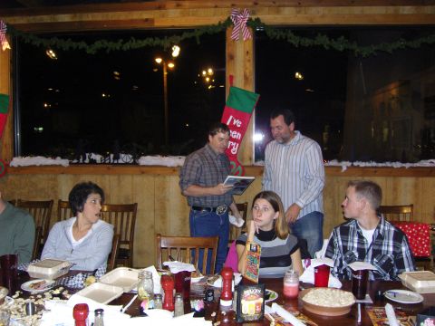 2007 Ford Mustang - A group of people are seated at a table, some looking at a man holding a booklet, during a holiday gathering.