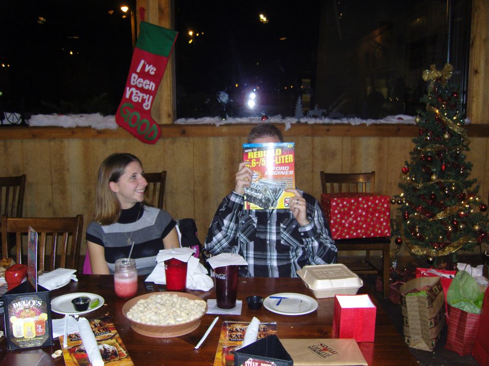People sitting at a table with a Christmas tree and decorations, one person holding a magazine about Ford engines