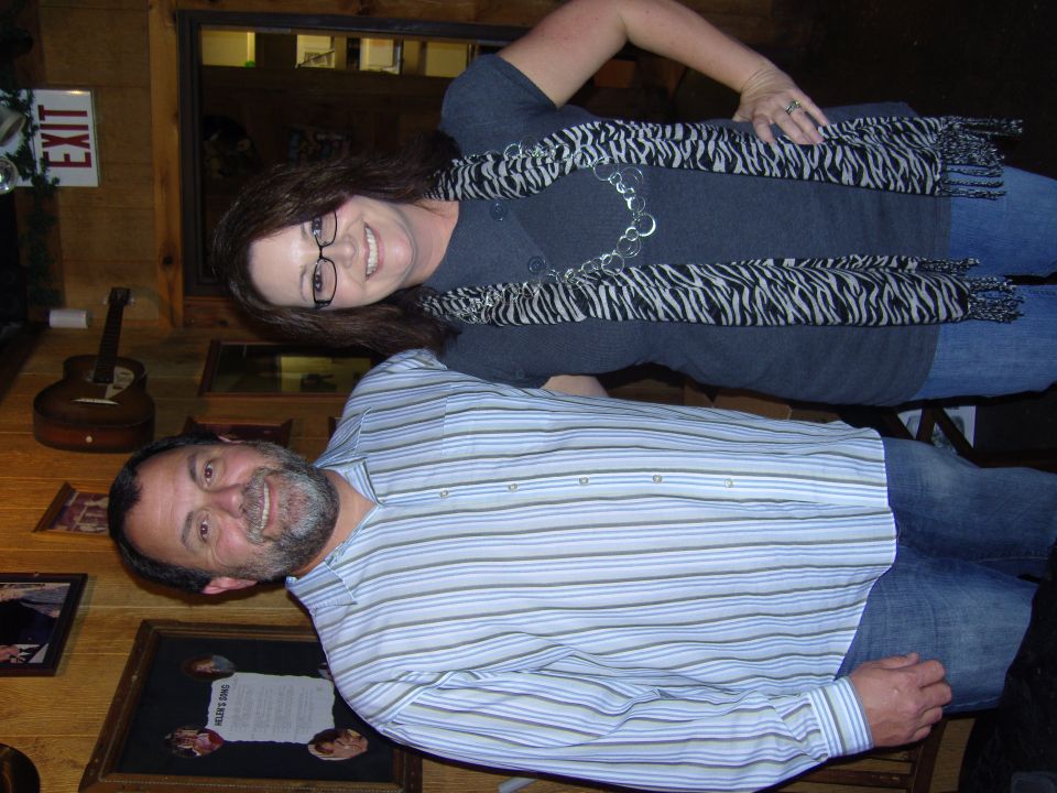 Two people posing for a photo in front of wood-paneled walls with an exit sign