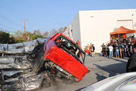 1978 Ford Bronco - A red off-road vehicle is overturned on a pile of crushed cars, with a crowd gathered to watch.