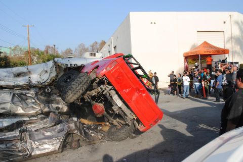1978 Ford Bronco - A red off-road vehicle rests precariously atop a stack of mangled cars, observed by onlookers.