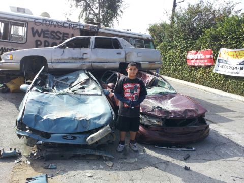 1978 Ford Bronco - A young boy poses with two severely damaged cars, with another vehicle and a bus stacked behind them.