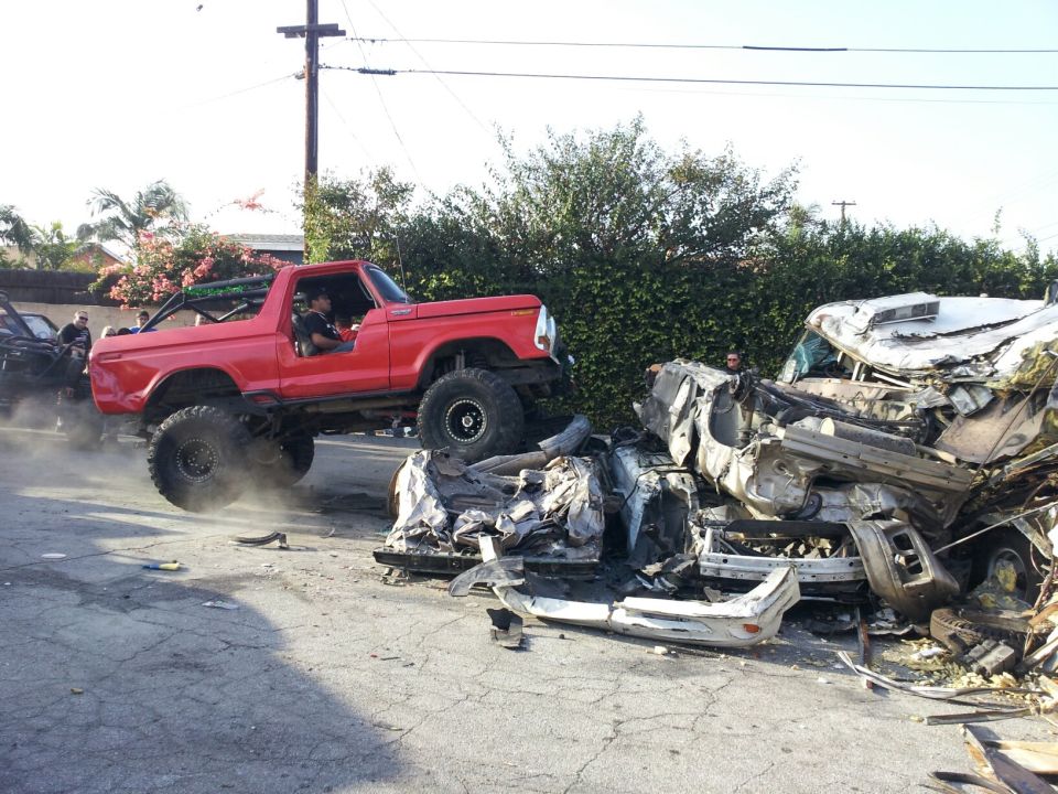 Red 1978 Ford Bronco with lifted suspension and large tires, mid-air over a severely damaged white car