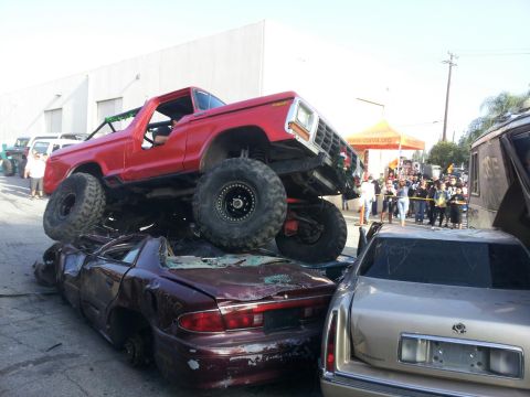 1978 Ford Bronco - A red 1978 Ford Bronco is shown crushing two cars, with spectators gathered behind a barrier.
