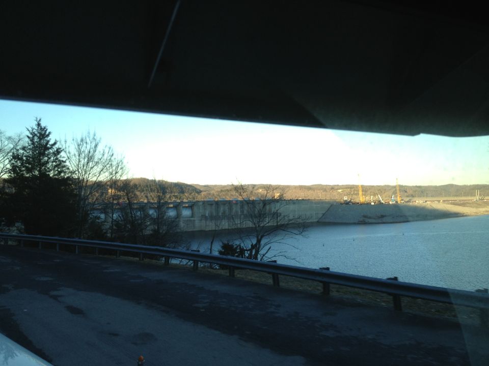 View of a large concrete dam and reservoir from a road, with trees and hills in the background
