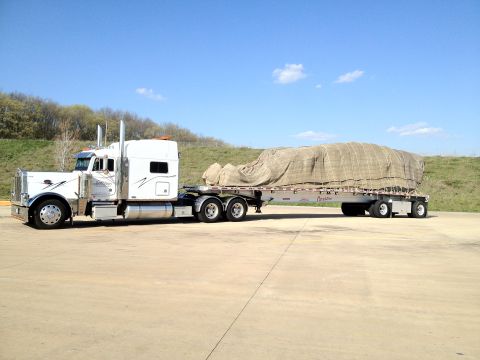 2003 Peterbilt 379 - A white semi-truck is parked on a concrete lot, hauling a large tarp-covered load on a flatbed trailer.