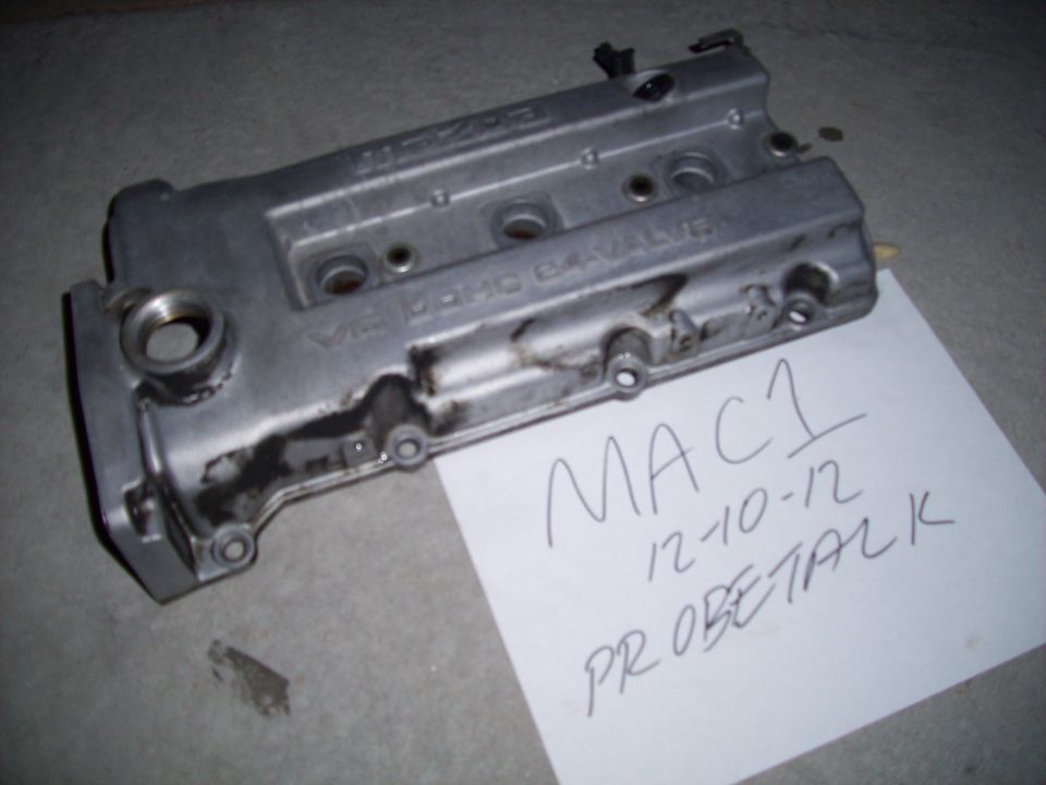 Silver engine valve cover with "V6 DOHC VALVE" text, resting on a concrete surface next to a handwritten note