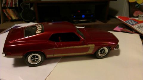 1970 Ford Mustang - A model car with a metallic red finish and gold racing stripes is displayed on a table.