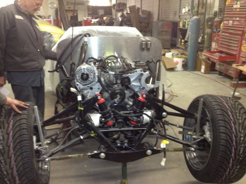 1975 Ford Ranchero - Front view of a bare hot-rod chassis in a shop, with a V8 mounted behind a tubular independent front suspension using red-bodied coilovers,…