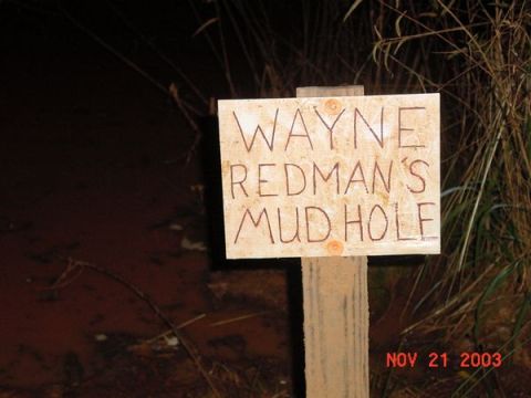 1989 Ford Bronco - A sign marks 'Wayne Redman's Mud Hole' next to a body of murky water and tall grass.