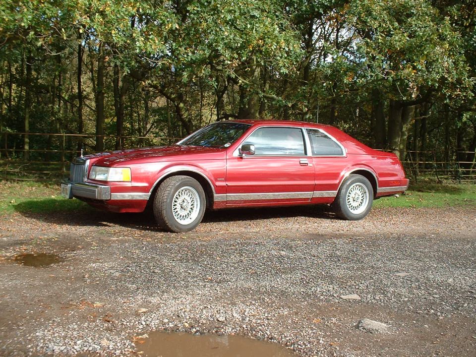 Red 1992 Lincoln Mark VII coupe parked on a gravel lot with trees in the background