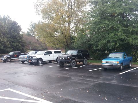 1986 Ford Bronco - A row of trucks and SUVs are parked in an outdoor lot on a cloudy day.