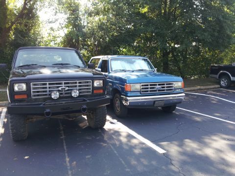 1986 Ford Bronco - Two Ford Broncos are parked in a lot, one black and one blue, with trees in the background.