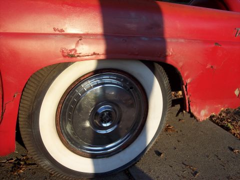 1956 Ford Thunderbird - Close-up of a rusty red 1956 Ford Thunderbird's side, showing a whitewall tire and chrome hubcap.