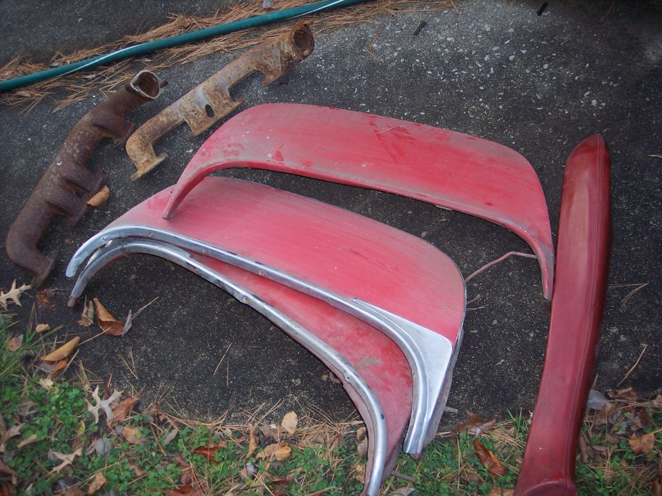 Several red car fender sections with chrome trim and two rusty exhaust manifolds on the ground