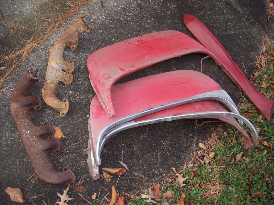 Two rusty exhaust manifolds and red car fender sections with chrome trim are on the ground