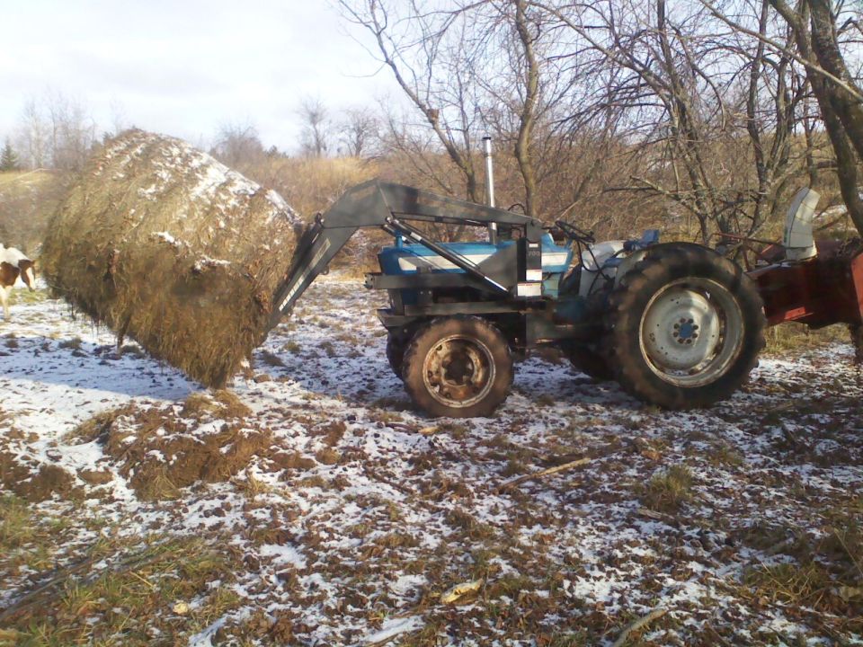 Blue tractor with a front loader lifting a large hay bale, with snow on the ground