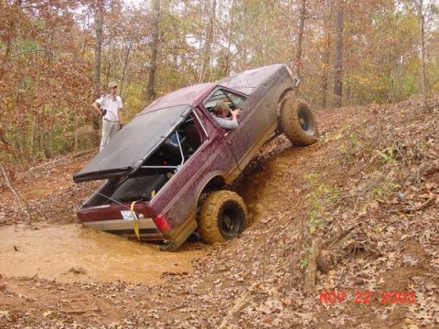 1989 Ford Bronco - A maroon Ford Bronco navigates a muddy incline in a wooded area, with one person inside and another standing nearby.