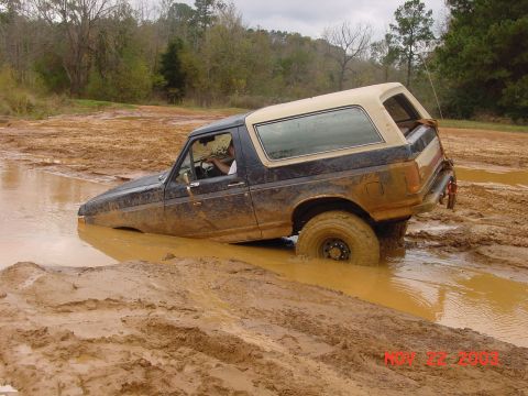 1989 Ford Bronco - A dark blue and tan Ford Bronco is stuck in deep mud and water, with trees in the background.