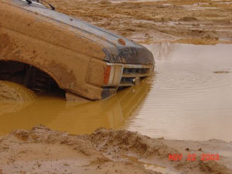 1989 Ford Bronco - A vehicle is stuck in a muddy puddle, with the front right wheel submerged in the brown water.