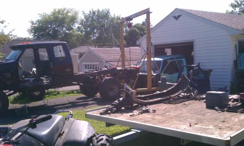 1994 Ford F-250 - A disassembled truck chassis sits in a yard with an engine hoist and various parts on a trailer.