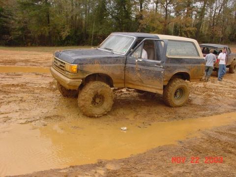 1989 Ford Bronco - A muddy 1989 Ford Bronco navigates a challenging off-road course with another vehicle visible in the background.
