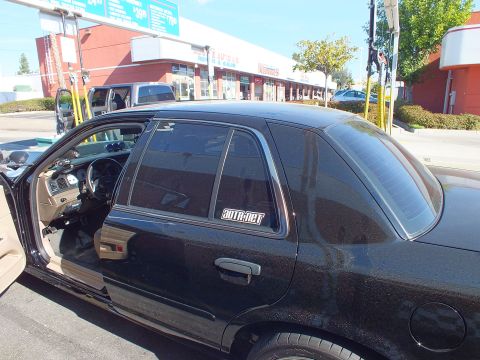 2003 Ford Crown Victoria - Side view of a black four-door sedan, front door open showing dashboard, parked outside red storefronts.
