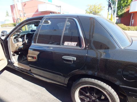 2003 Ford Crown Victoria - Passenger profile of a black four-door sedan with door ajar, tinted windows and dark alloy rear wheel.