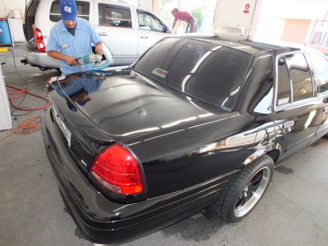 2003 Ford Crown Victoria - Worker buffs the rear deck of a black four-door sedan with chrome-lip wheels inside a detail bay.