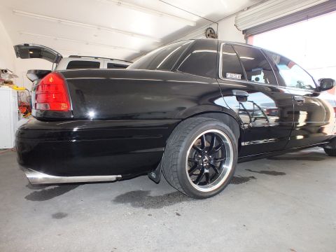 2003 Ford Crown Victoria - Rear quarter of a black sedan, trunk lid raised, low-profile tires on black alloys inside a shop.