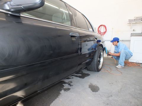 2003 Ford Crown Victoria - Detailer kneels at the rear wheel of a black sedan, coiled red air hose on the bay floor behind.