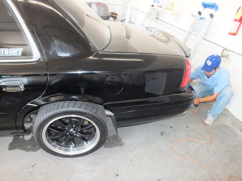 2003 Ford Crown Victoria - Black sedan rear quarter and dark alloy wheel while a worker in blue services the bumper