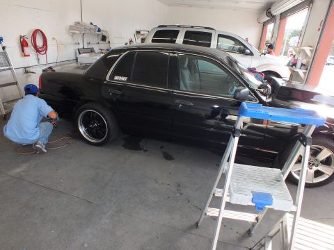 2003 Ford Crown Victoria - Side view of lowered black sedan in shop bay, technician working at the front wheel